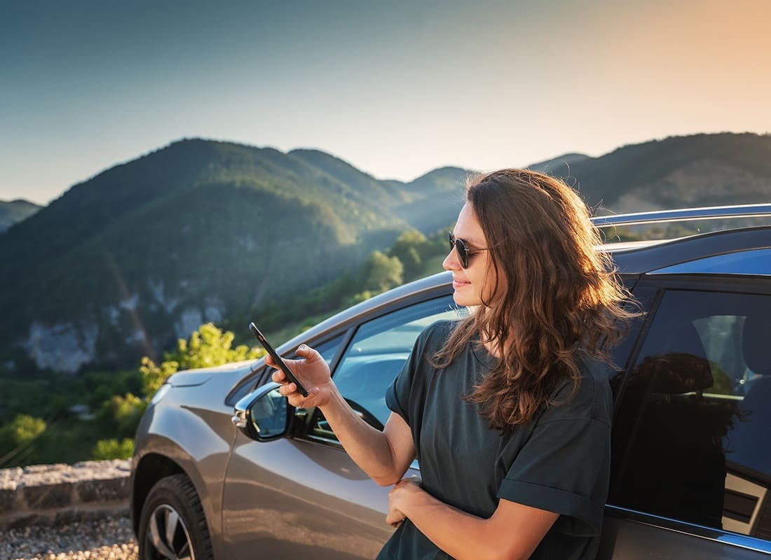 Service Center - Portrait of a Young Woman Holding a Phone in her Hands While Leaning Against her Car Parked at a Lookout Point with Views of the Mountains